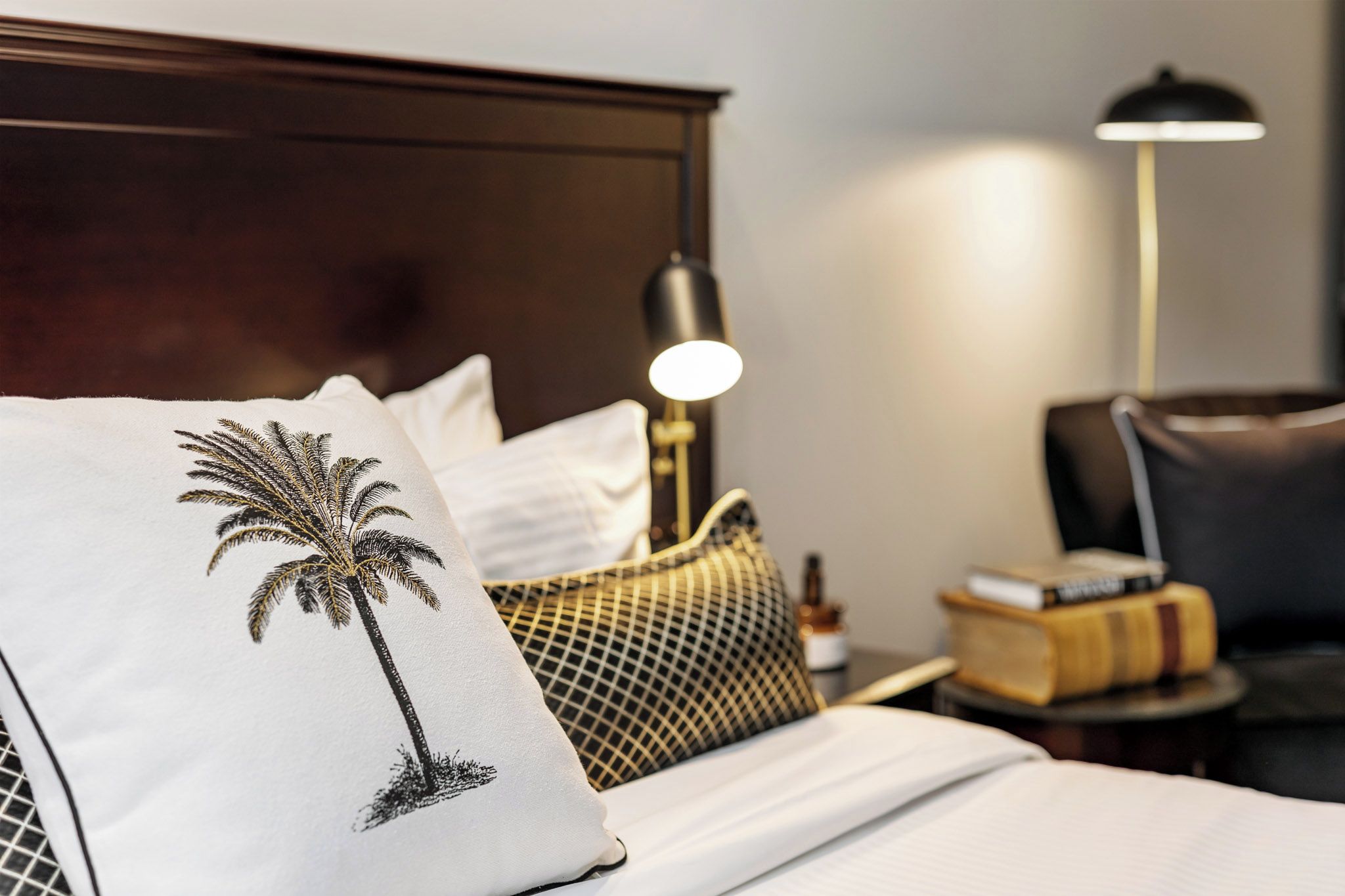 A neatly made bed with a pillow featuring a palm tree design, a bedside lamp, a checkered throw pillow, and a small bedside table with a book and a bottle.