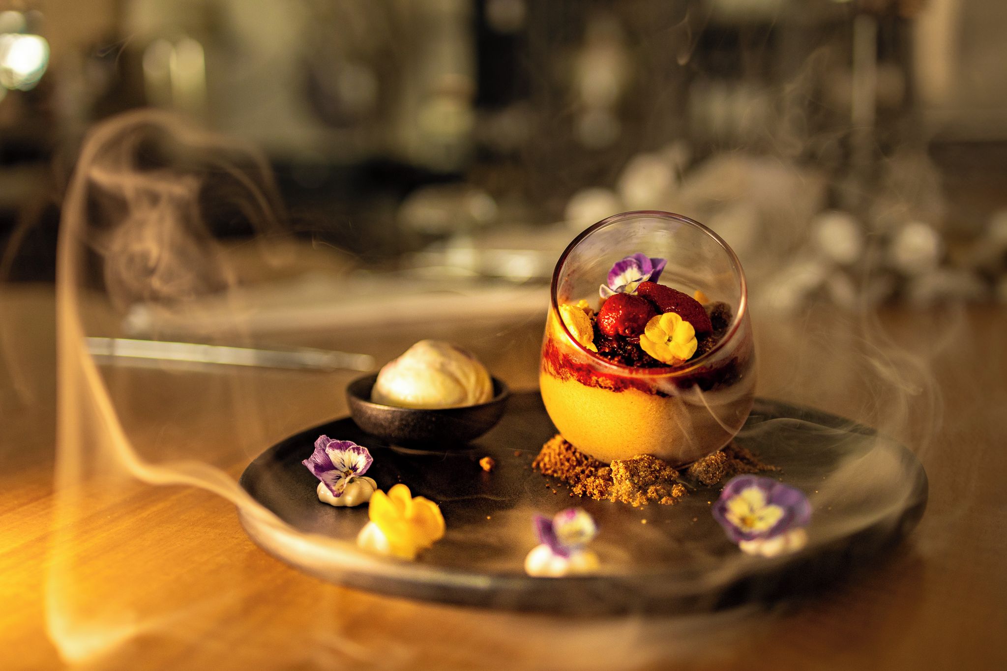 Un postre con una cúpula de vidrio sobre un plato, acompañado de un recipiente pequeño de crema y flores comestibles esparcidas.
