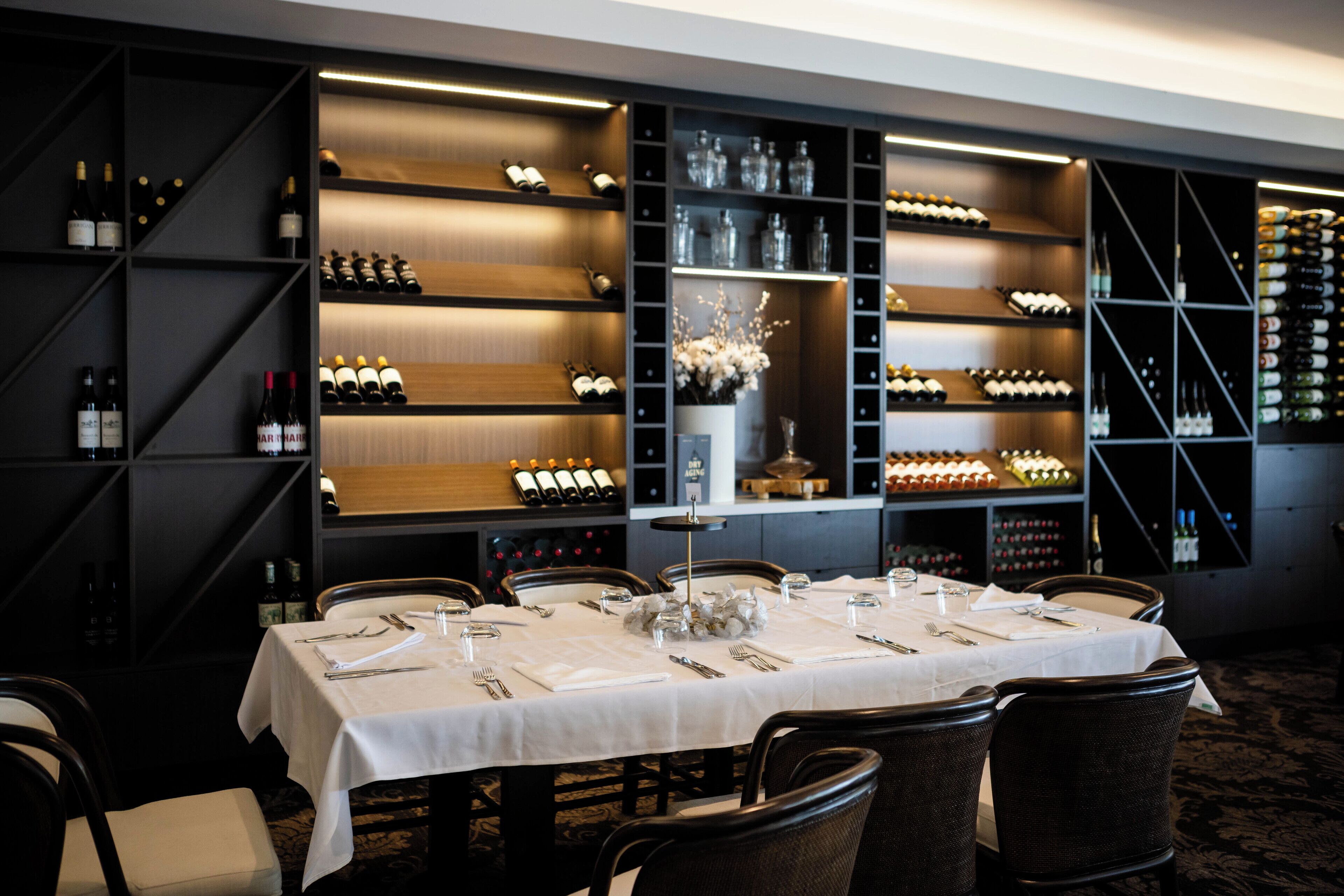 Un comedor con una mesa dispuesta para seis personas, rodeada de sillas, y estanterías llenas de botellas de vino y copas al fondo.
