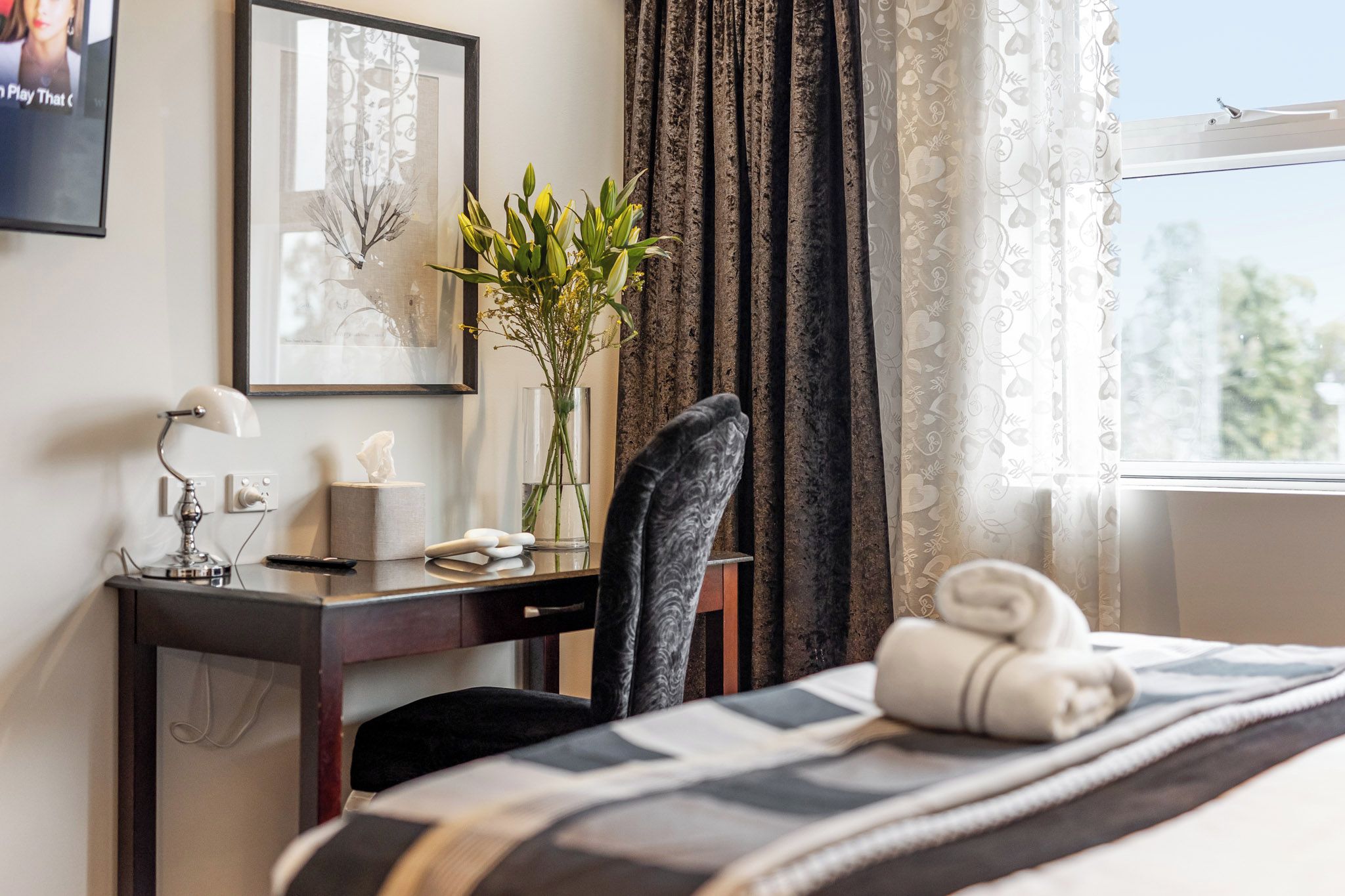 Una habitación de hotel con una cama, un escritorio con lámpara, una silla, un florero con flores, un cuadro enmarcado y una ventana con cortinas.