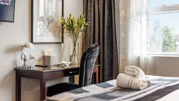 Una habitación de hotel con una cama, un escritorio con lámpara, una silla, un florero con flores, un cuadro enmarcado y una ventana con cortinas.