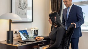 A woman seated at a desk with a laptop, a remote control, and a lamp. A man stands beside her, holding a jacket. - International Hotel Wagga Wagga (East Wagga Wagga)