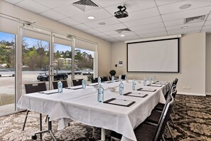 Un salón de conferencias con una larga mesa, sillas, botellas de agua y una pantalla para proyector.