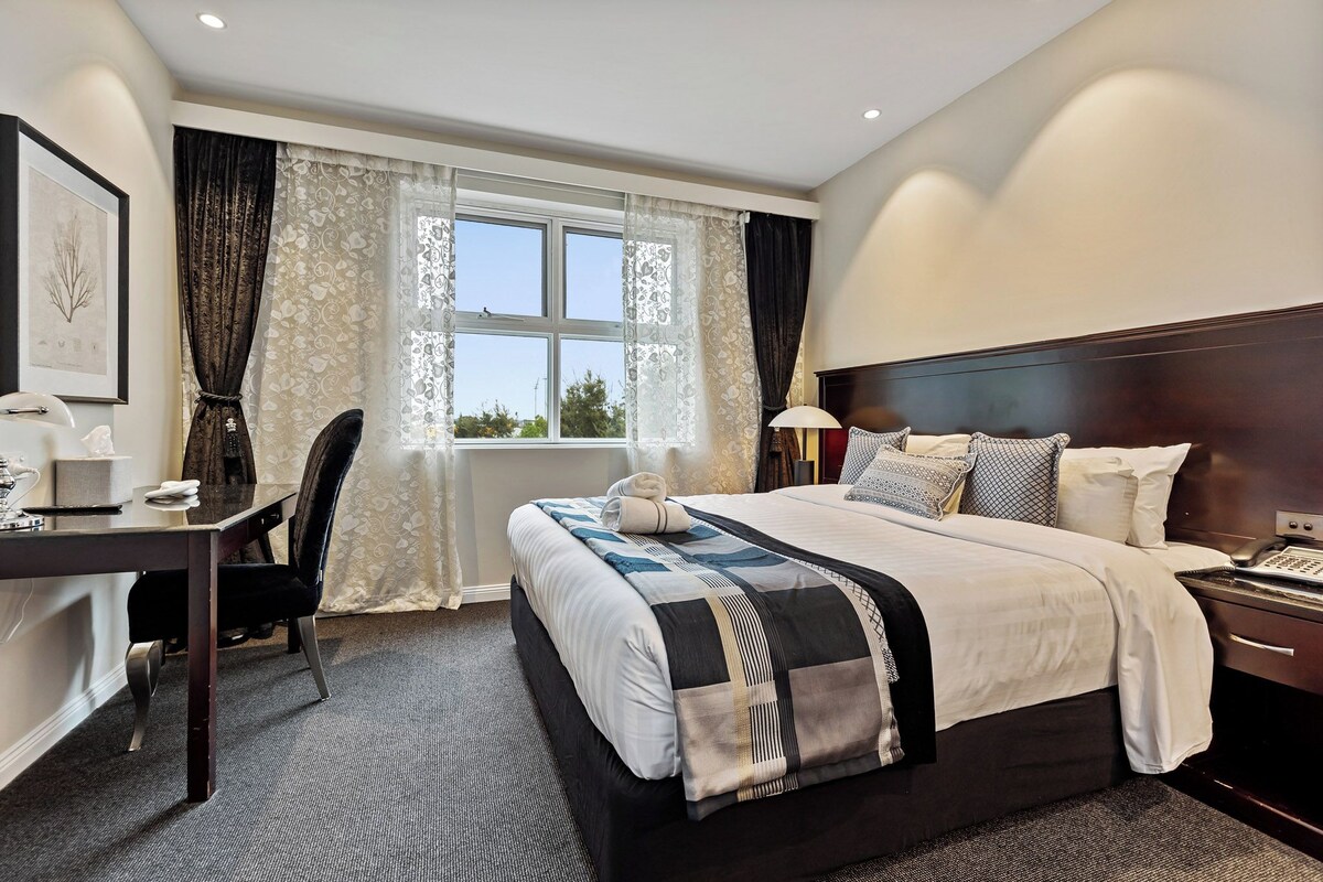 A hotel room with a large bed, a desk with a chair, a bedside table with a lamp, and a window with curtains.