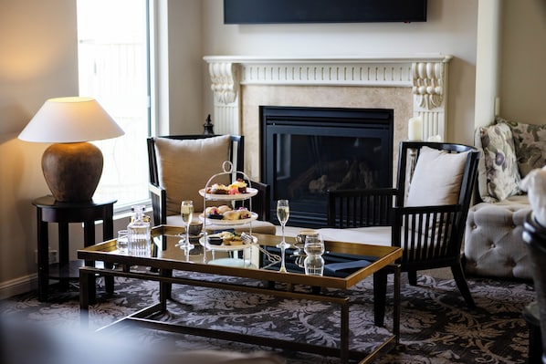 A cozy living room with a fireplace, a coffee table with a tray of pastries, a lamp, and a television mounted on the wall.