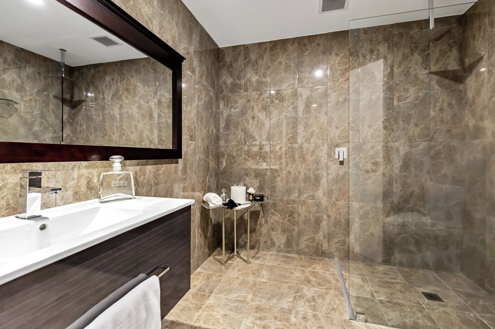 A modern bathroom with a large mirror, a white sink, a dark vanity, a shower area, and a small table with toiletries.