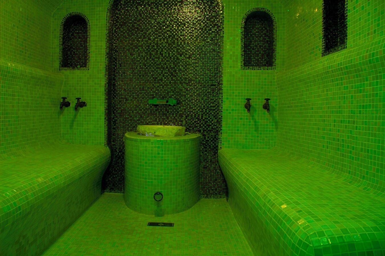 Een badkamer met groene tegels, een centraal geplaatst wastafel en twee bankjes.