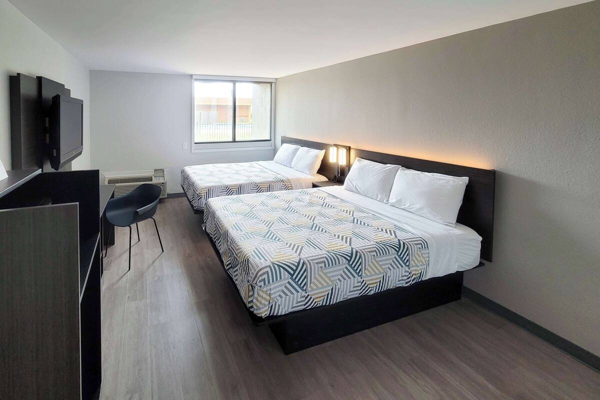 A hotel room with two beds, a desk, a chair, a TV, and a window with natural light.