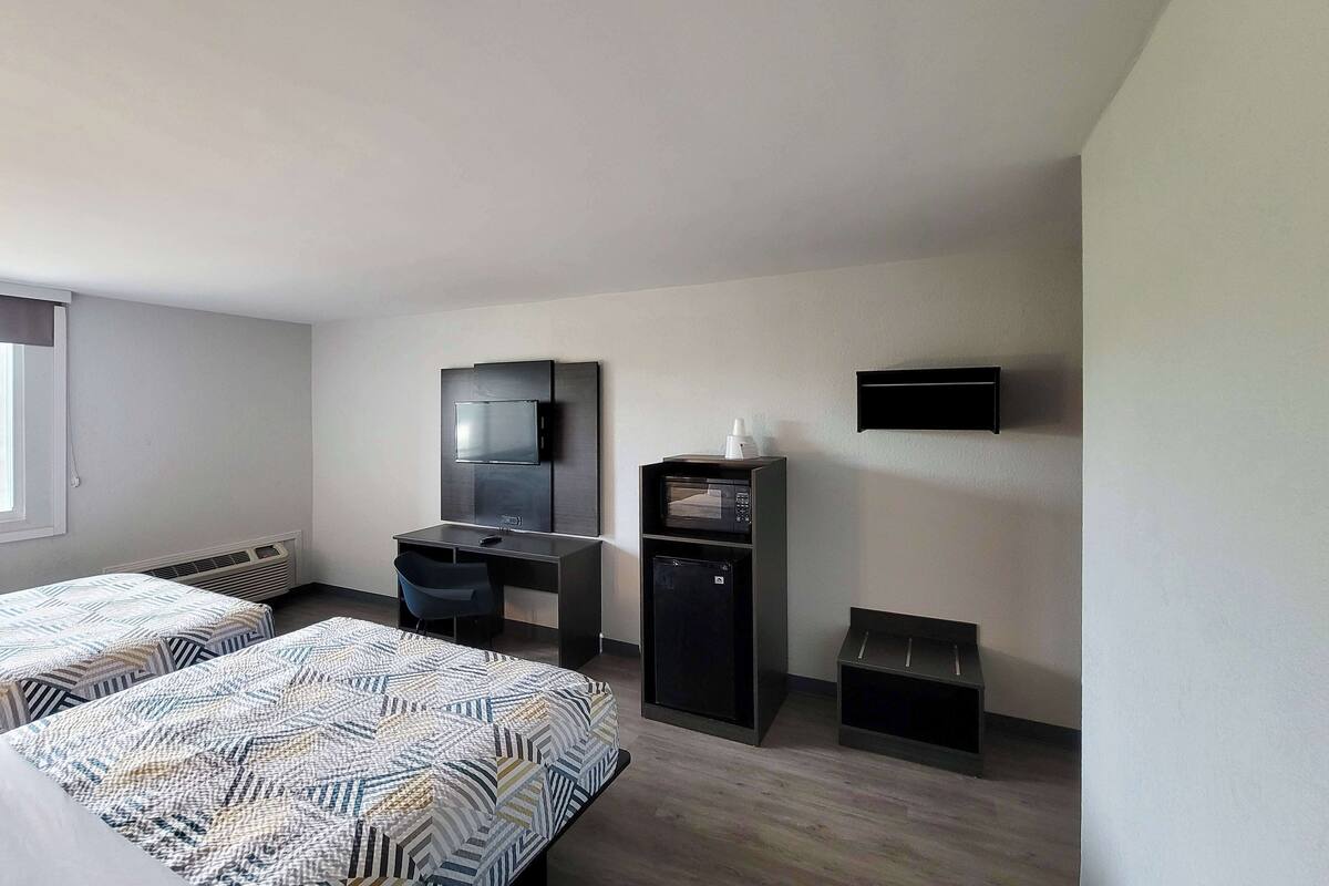 A compact hotel room with two beds, a desk, a microwave, and a TV mounted on the wall.
