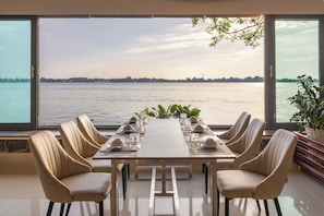 A dining table with six chairs, set for a meal, overlooking a body of water with a distant shoreline. - Van Phat Riverside Hotel (Can Tho)
