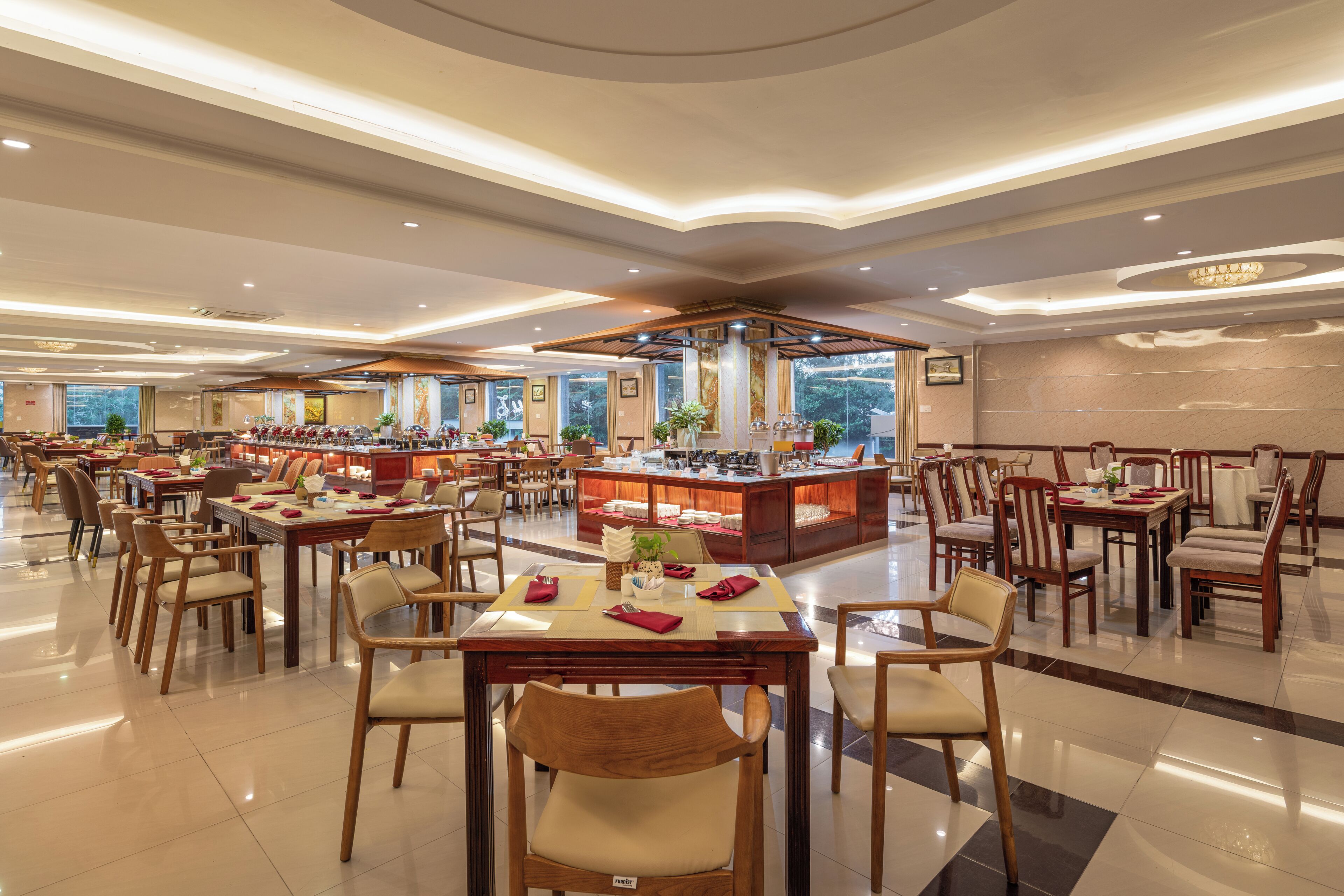 A spacious hotel dining area with multiple tables and chairs, a central buffet station, and large windows providing natural light.