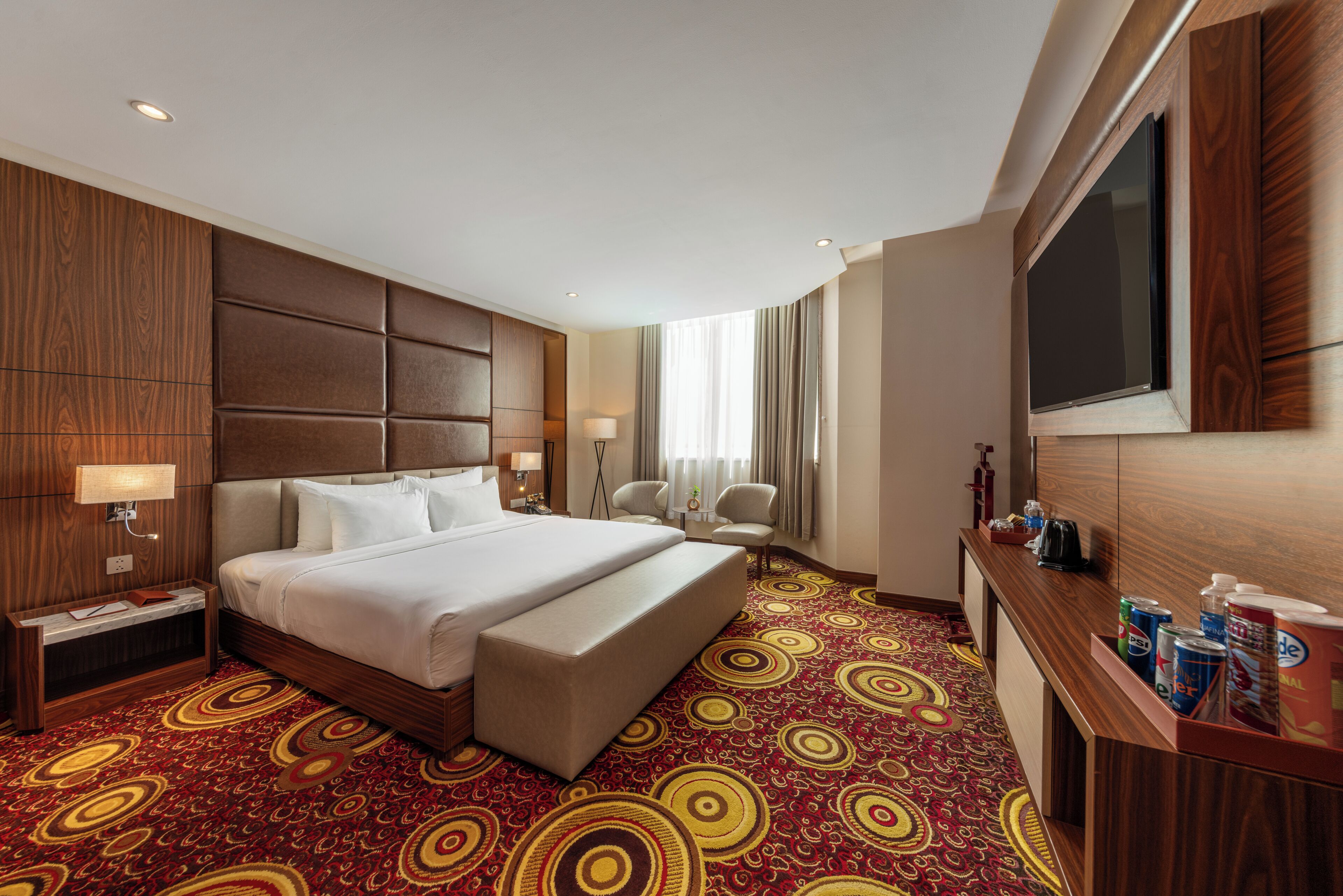 A hotel room with a large bed, a desk with a flat-screen TV, a minibar, and a patterned carpet.
