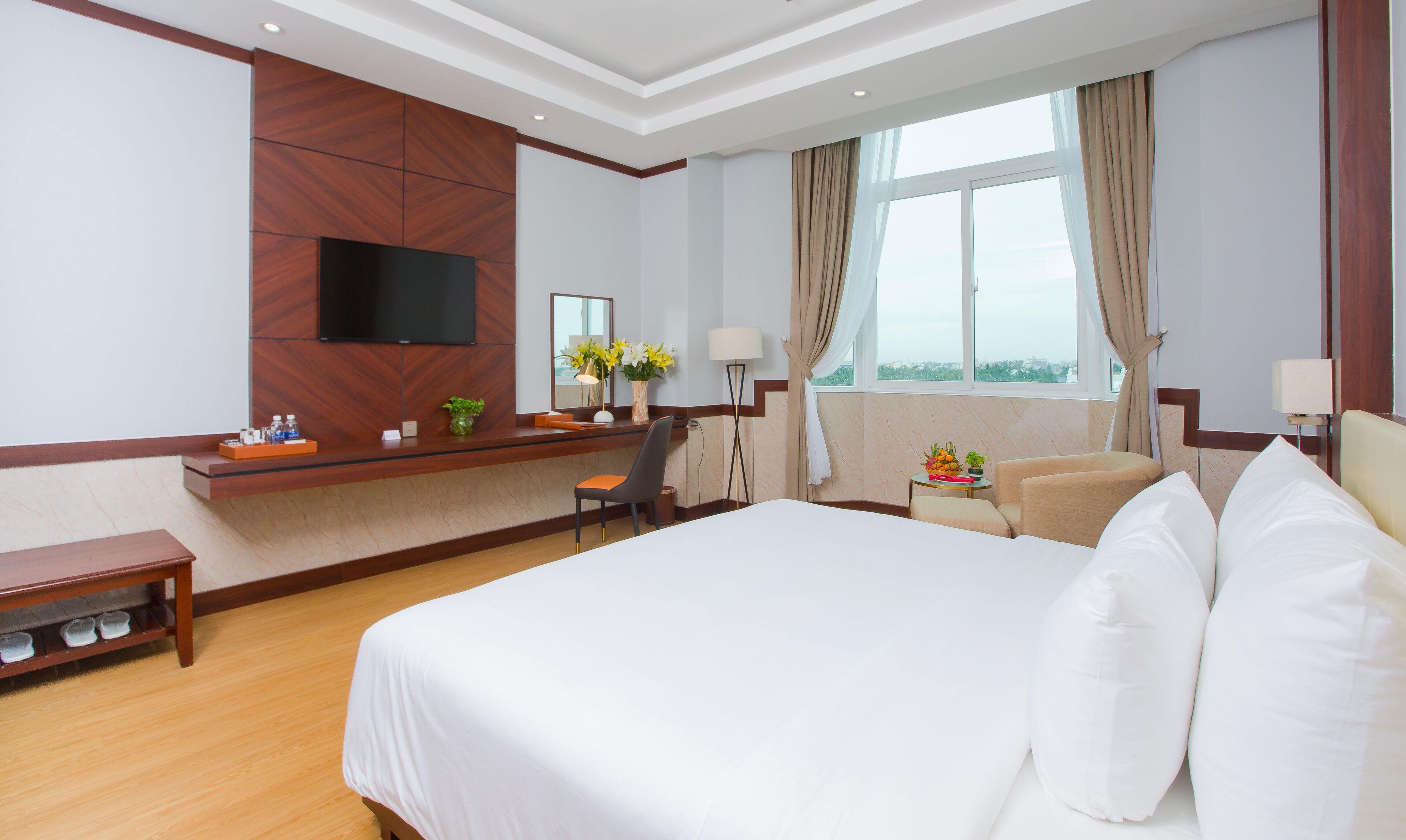 A hotel room with a large bed, a desk with a flat-screen TV, a chair, a small table with a lamp, and a window with curtains.