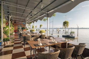 An outdoor dining area with checkered flooring, tables, chairs, hanging plants, and a view of the water. - Van Phat Riverside Hotel (Can Tho)