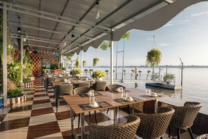 An outdoor dining area with checkered flooring, tables, chairs, hanging plants, and a view of the water.