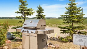 A portable gas grill labeled "GENESS" in a grassy area with a sign that reads "GUEST BARBEQUE OPEN ALL YEAR ROUND."