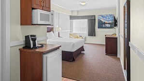 A hotel room with a bed, a desk with a coffee maker, a microwave, a small refrigerator, a chair, a window with curtains, and a painting on the wall.