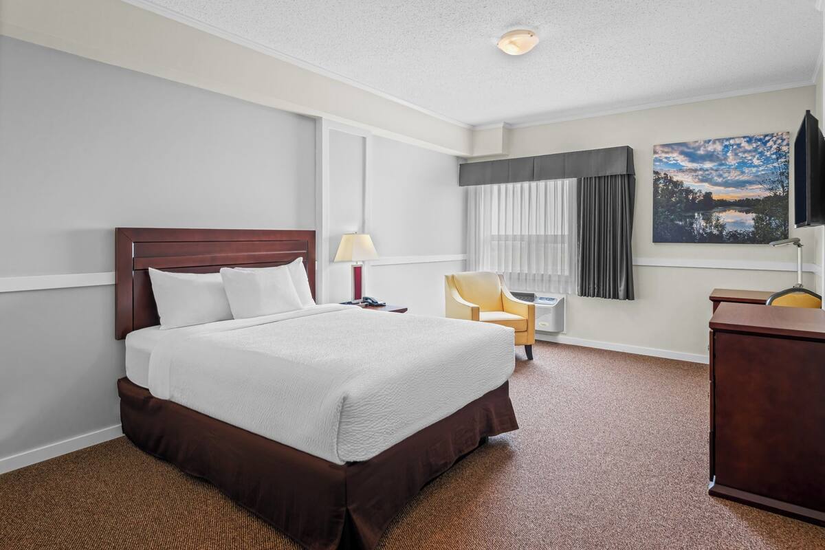 Une chambre d’hôtel avec un grand lit, un bureau, une chaise, une lampe, une télévision et une fenêtre avec des rideaux.