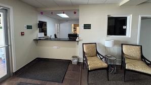 A reception area with a front desk, two chairs, a small table, a waste bin, and a check-in/out sign.