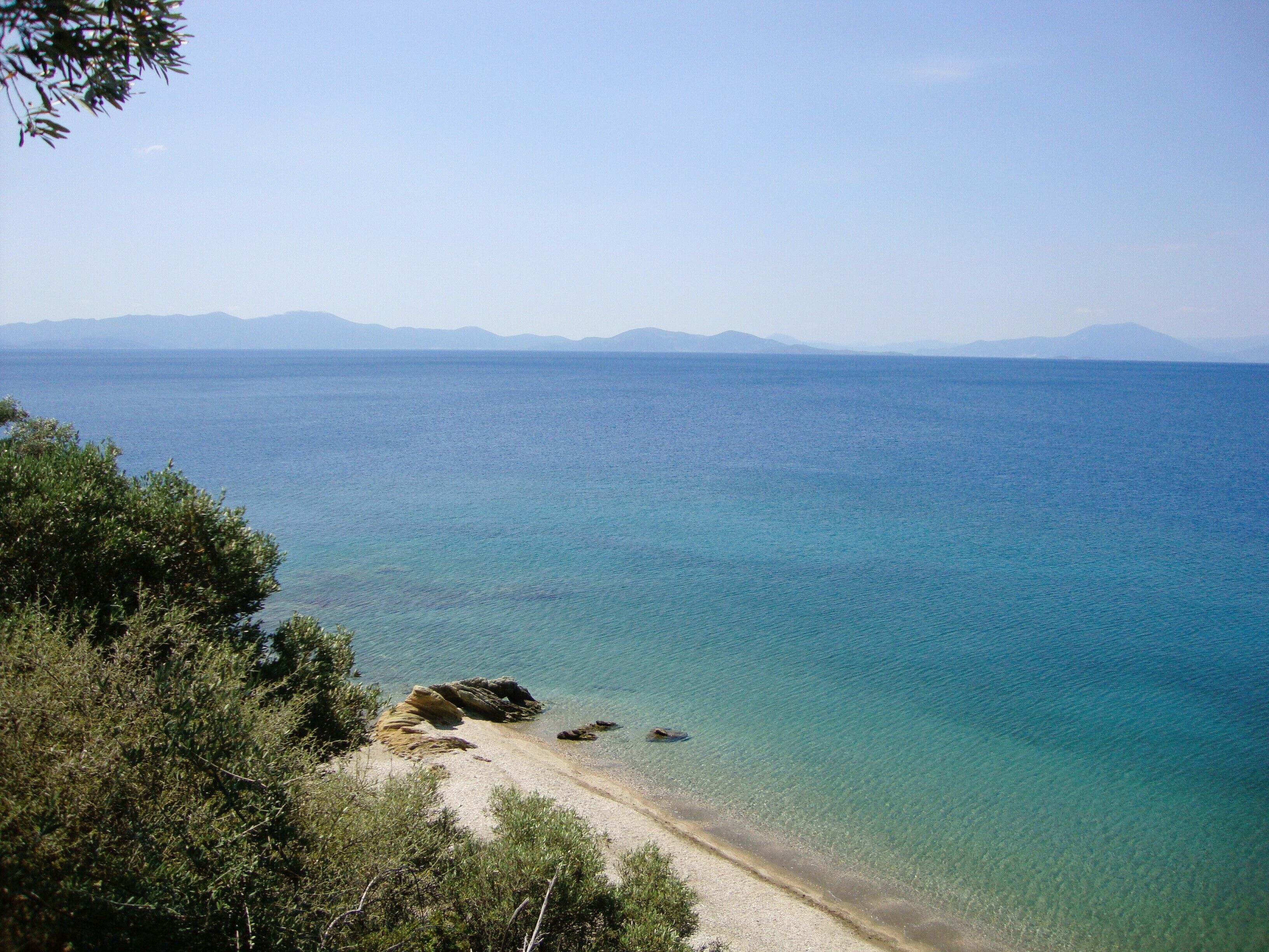 A coastal landscape with clear blue waters, a sandy beach, and distant mountains.