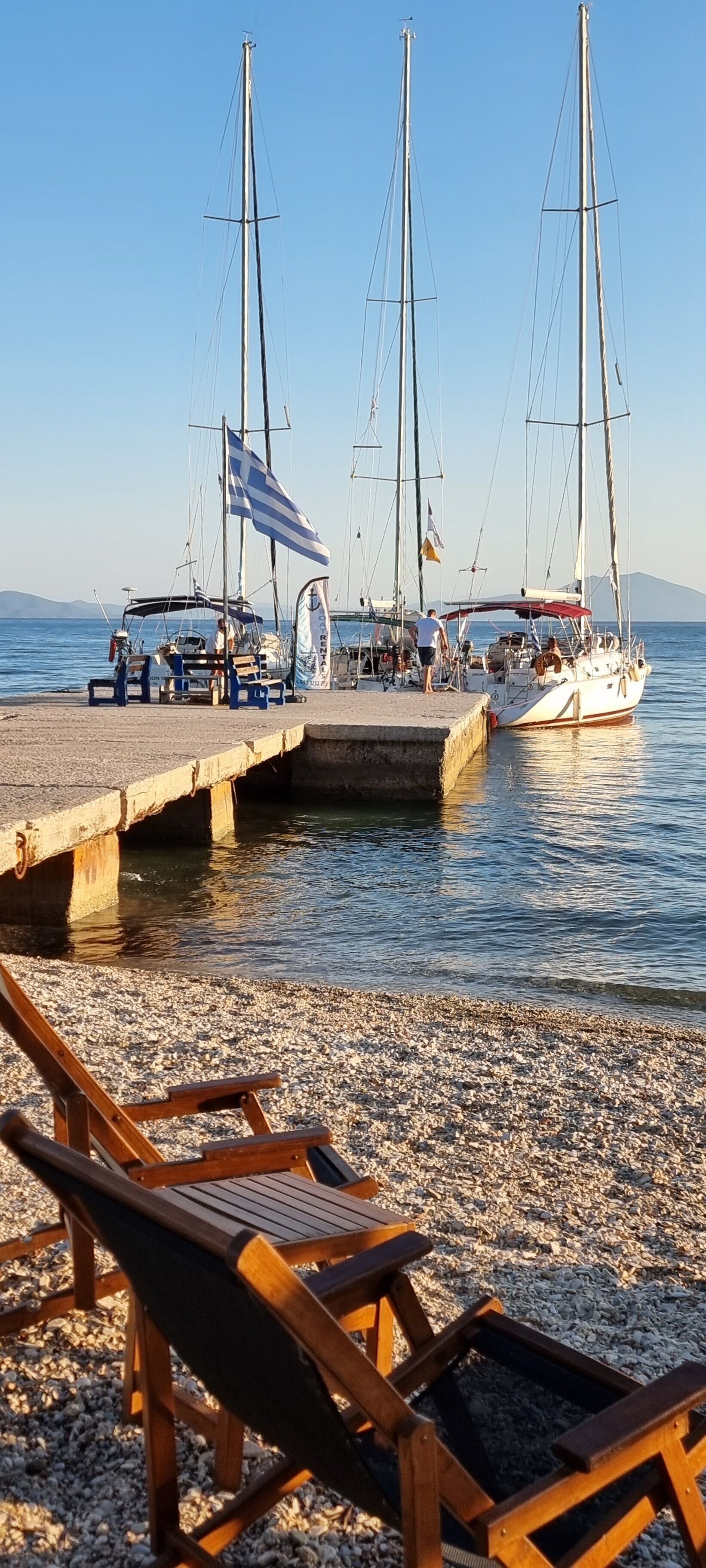 A pebbled beach with wooden lounge chairs and a dock with sailboats.