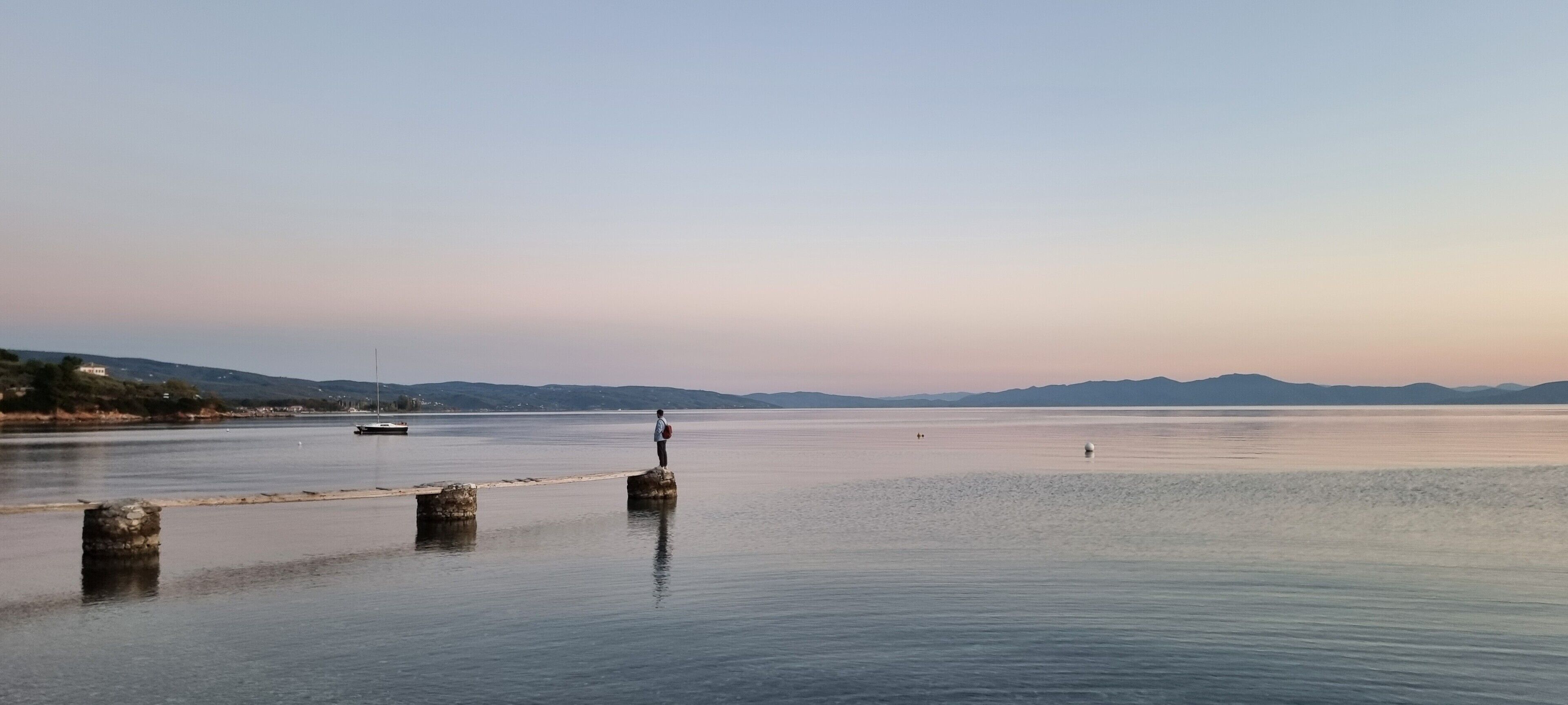 Un lac paisible, avec une personne debout sur une jetée, des bateaux à l’horizon et un paysage vallonné en arrière-plan.
