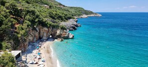 A coastal beach with clear turquoise waters, rocky cliffs, and a few buildings on the hillside.