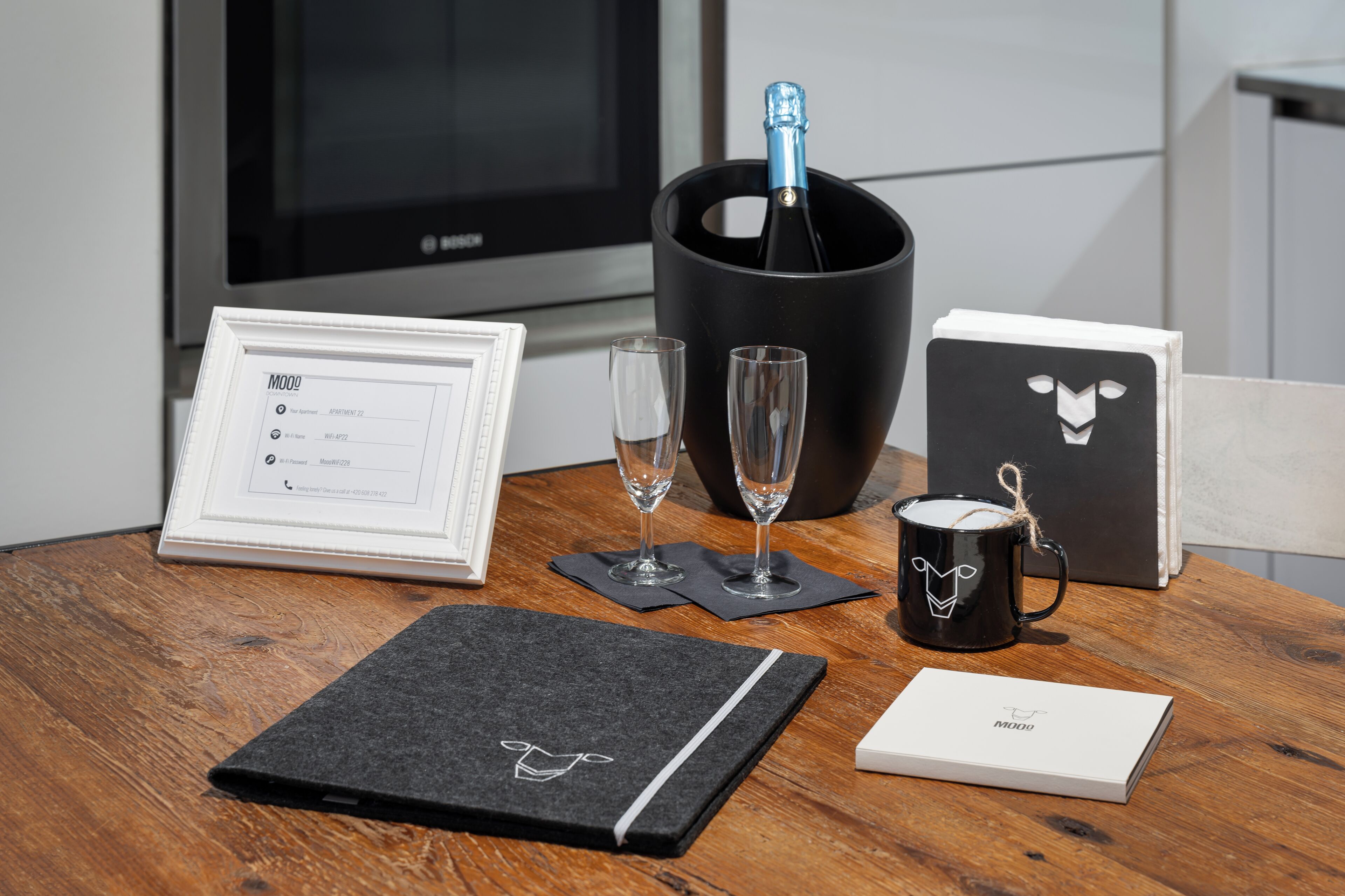 A wooden table with a framed menu, two glasses, a bottle of champagne in a black bucket, a black mug, and a black notebook with a white logo.