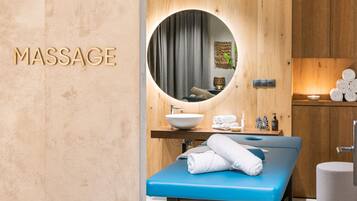 A massage room with a wooden wall, a round mirror, a blue massage table, and a sink with towels.