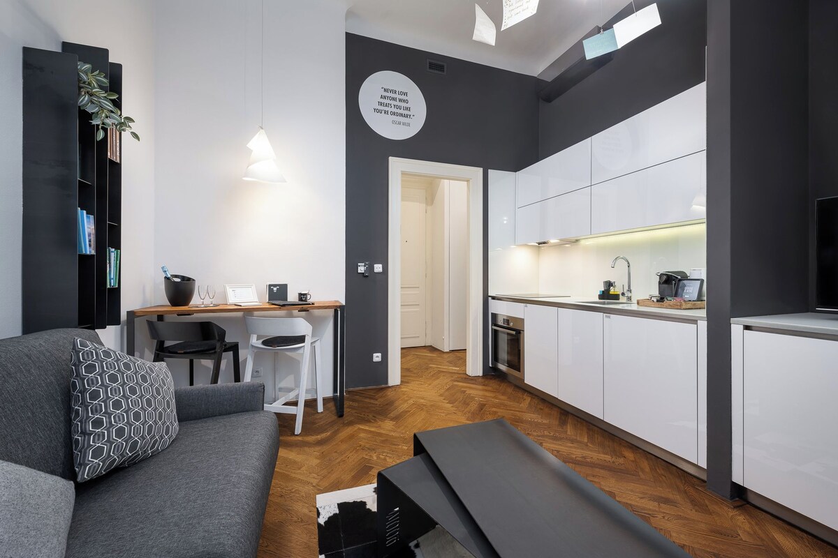 A modern apartment with a grey sofa, a dining area, a kitchen with white cabinets, and a black bookshelf.