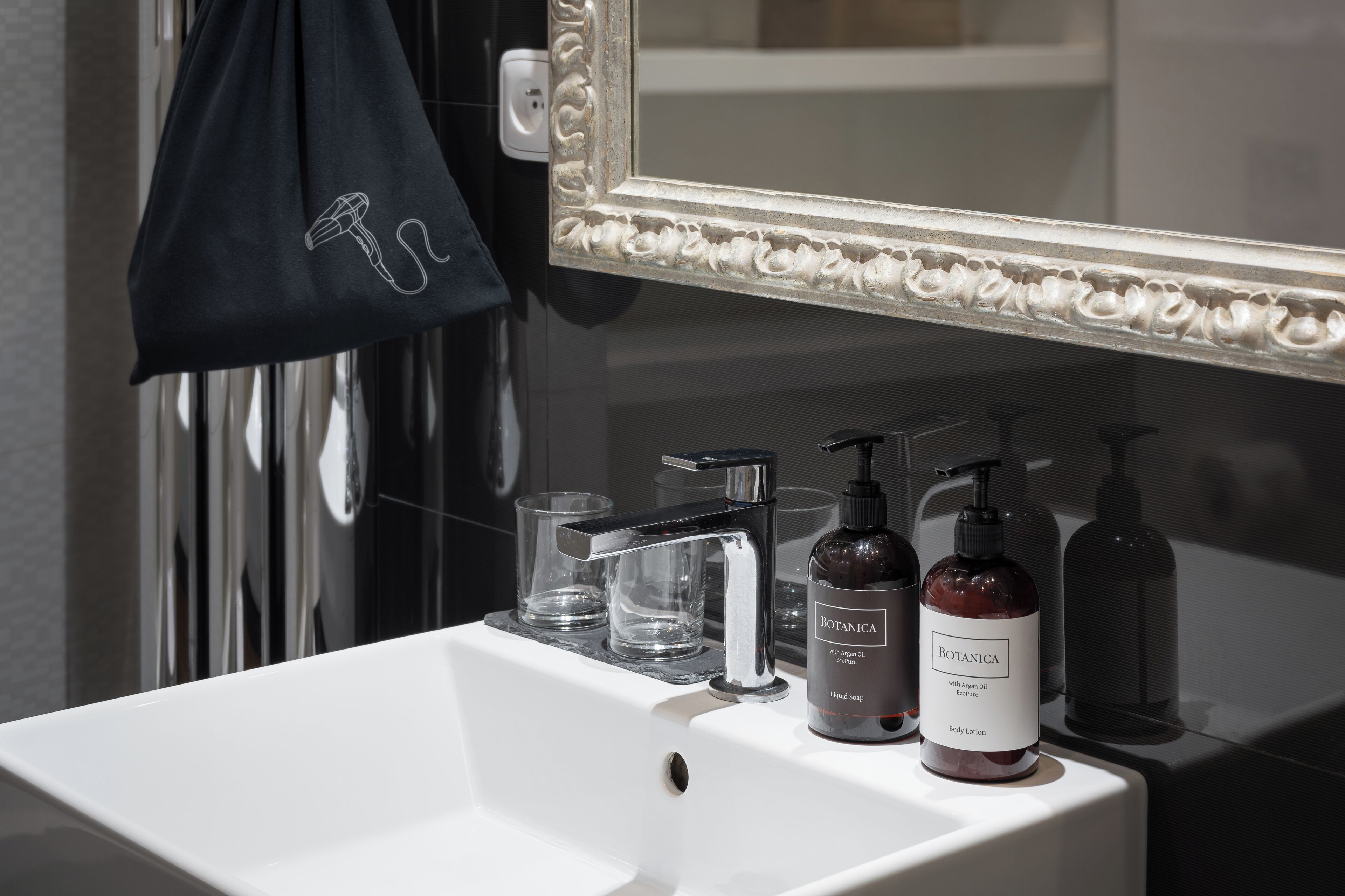 Casa de banho com lavatório branco, dois copos de vidro transparente, toalha preta e dois frascos de loção corporal Botanica.