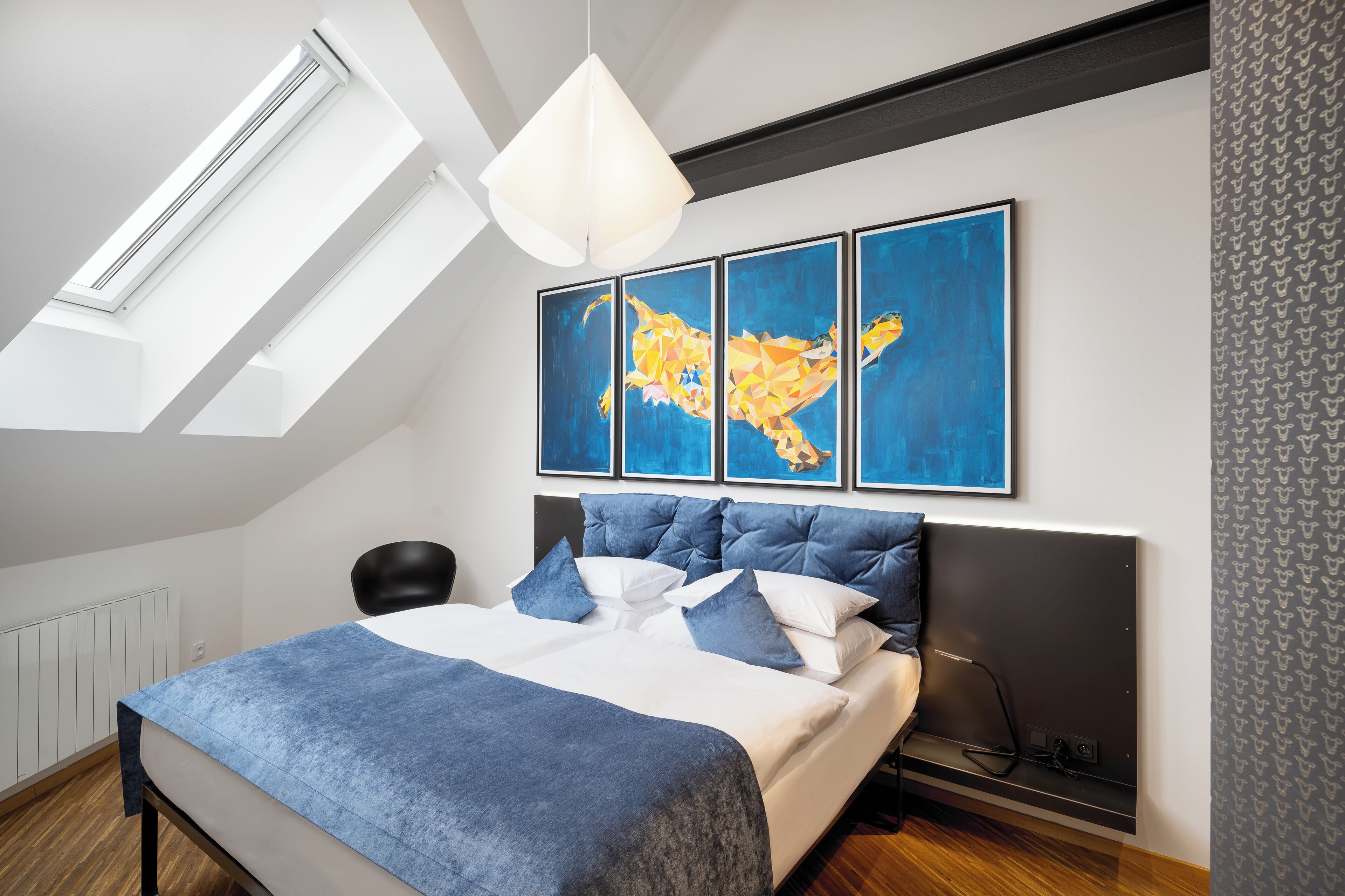 Quarto moderno com claraboia, cama com roupa de cama azul e três quadros abstratos na parede.