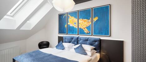 Quarto moderno com claraboia, cama com roupa de cama azul e três quadros abstratos na parede.