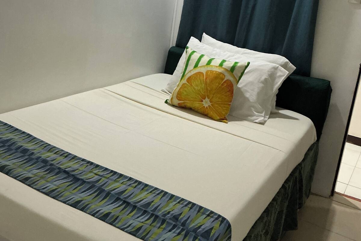 Ein Einzelbett mit einem gemusterten Bettbezug, ein Kissen mit einem Orangenscheiben-Motiv und ein blauer Vorhang.