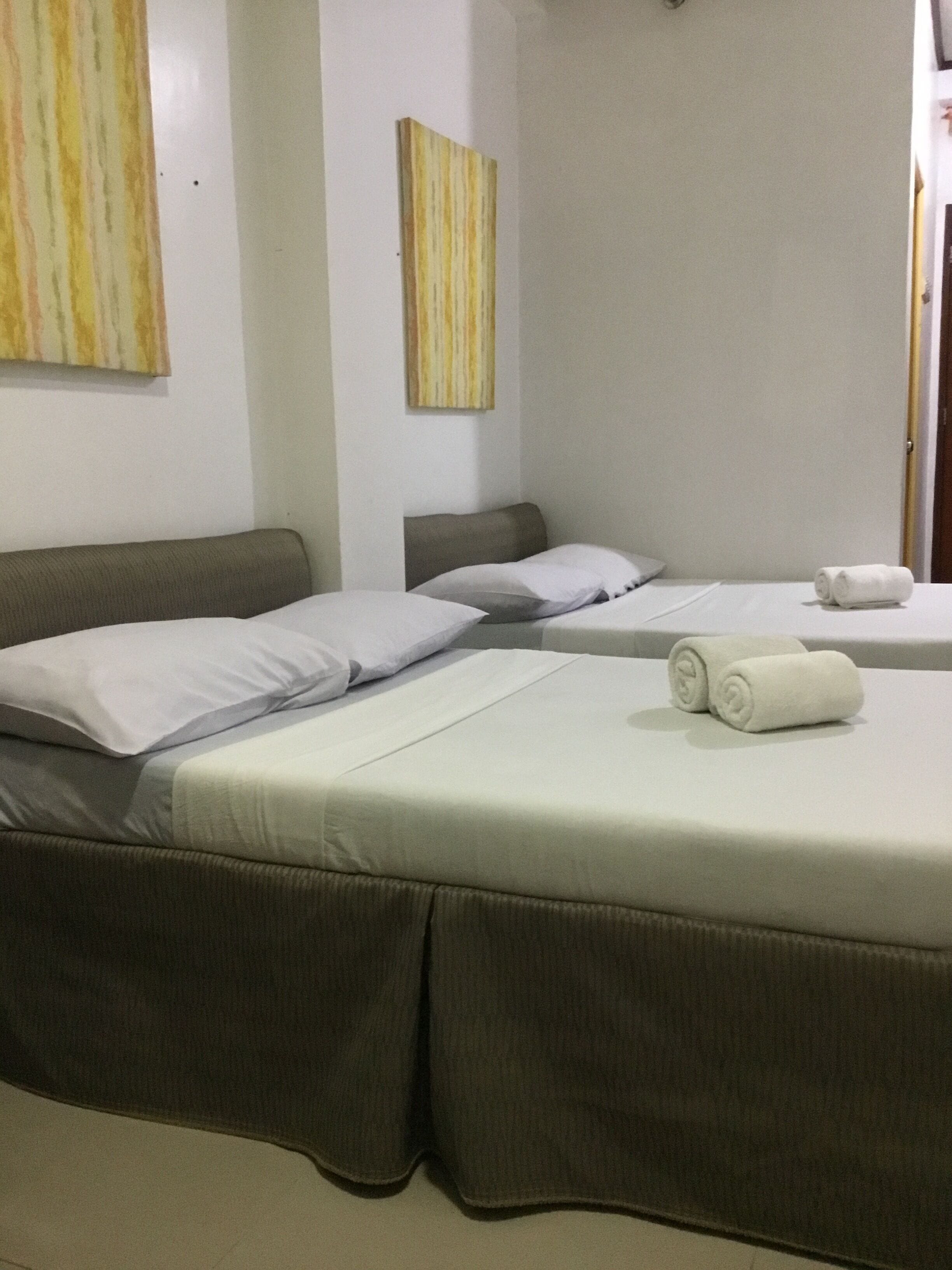 Une chambre d’hôtel avec deux lits, chacun doté d’une tête de lit grise et de linge de lit blanc. Deux tableaux sont accrochés aux murs, un au-dessus de chaque lit. Une serviette roulée est posée sur le lit, au premier plan.