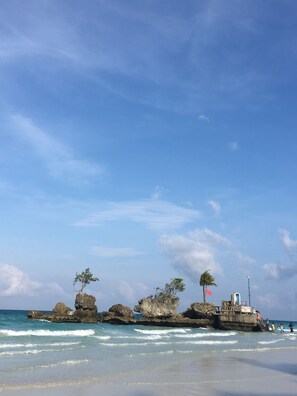 Una playa con afloramientos rocosos, cielo azul despejado y mar tranquilo.