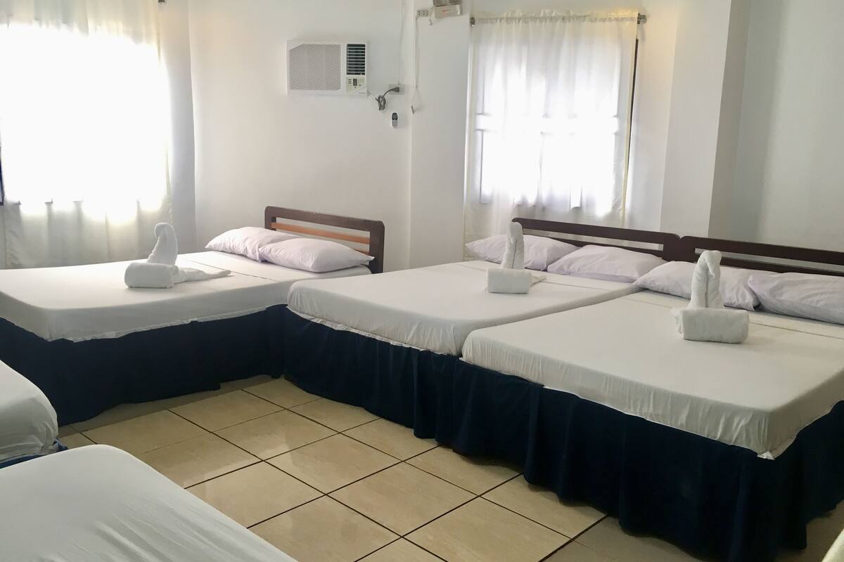 Ein Hotelzimmer mit zwei Betten, jeweils mit einem Holz-Kopfschild, weißen Laken und einem dunkelblauen Bettrock. Es gibt ein Fenster mit durchscheinenden Vorhängen, eine Klimaanlage an der Wand und einen wandmontierten Lichtschalter.