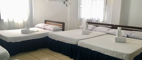 A hotel room with two beds, each with a wooden headboard, white linens, and a dark blue skirt. There is a window with sheer curtains, an air conditioning unit on the wall, and a wall-mounted light switch.