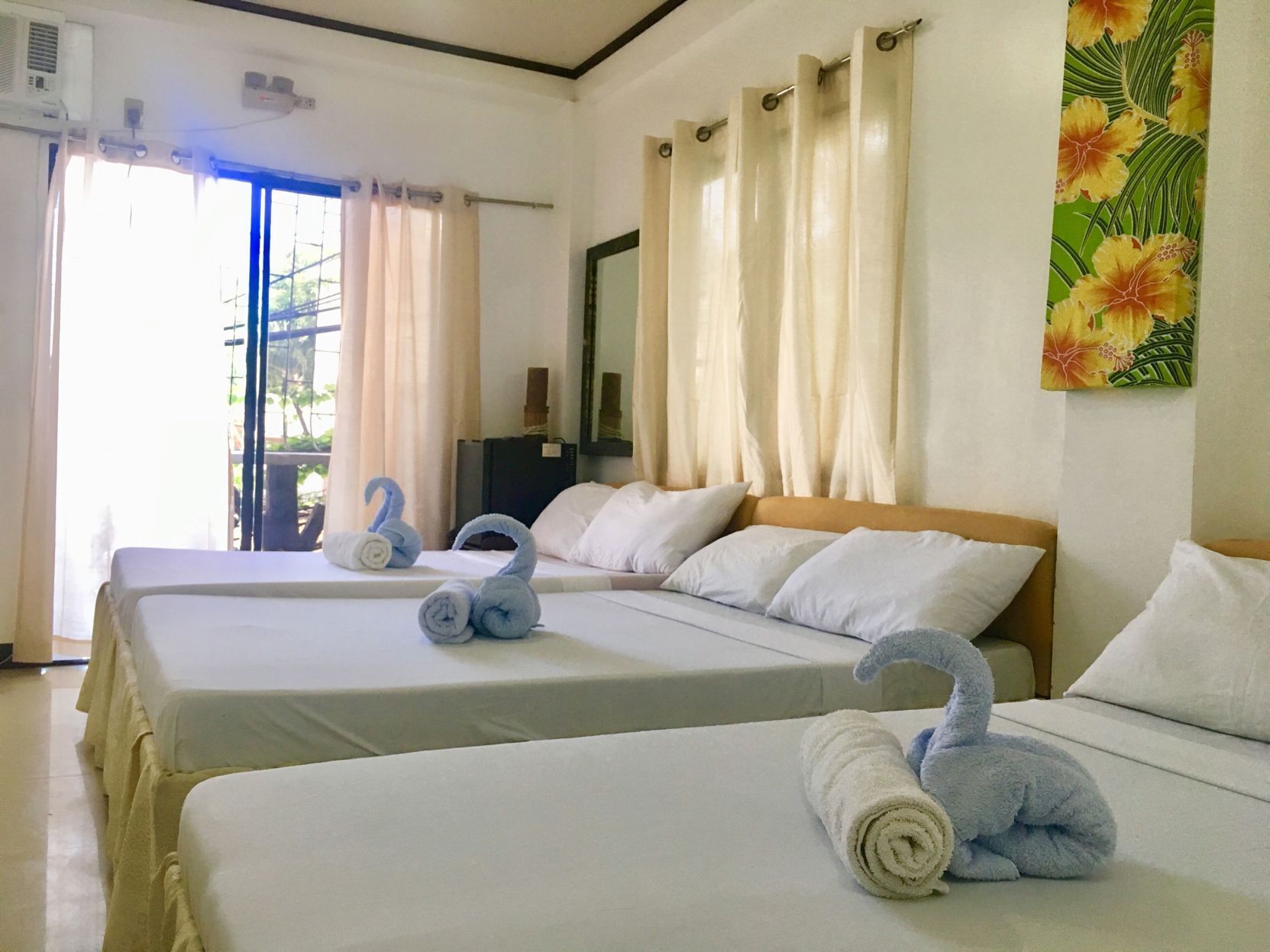 A hotel room with two beds, a large window with curtains, a wall-mounted air conditioner, and a painting.
