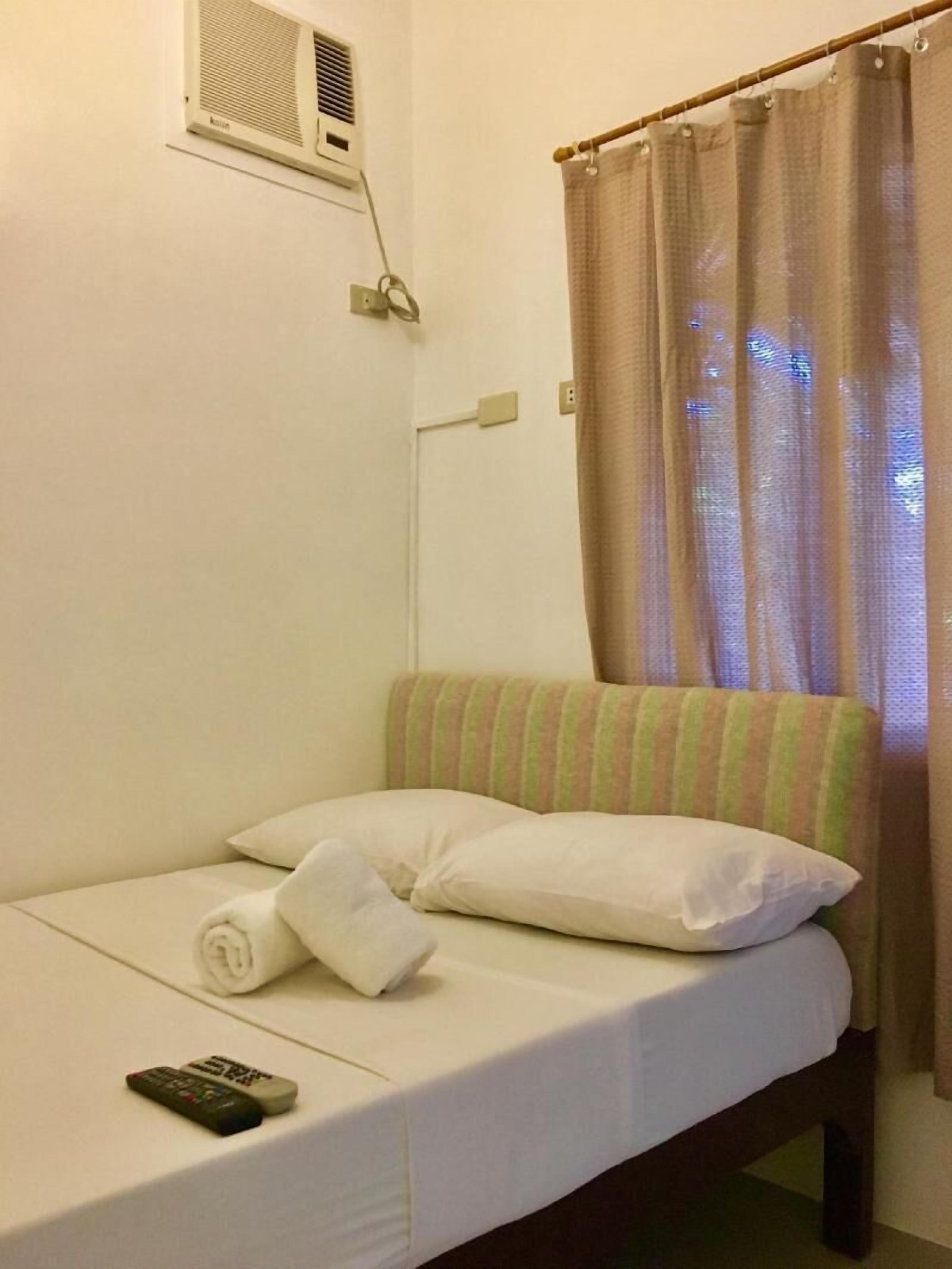 A single bed with a pillow and a rolled towel, a remote control, a wall-mounted air conditioner, and a curtain.