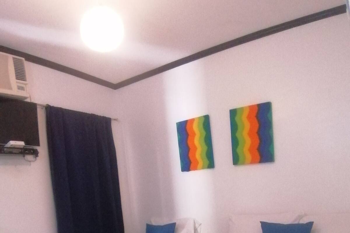 Ein Hotelzimmer mit zwei Einzelbetten, jeweils mit blauer Bettdecke und weißen Kissen. Es gibt ein Fenster mit dunklem Vorhang, eine Klimaanlage an der Wand und zwei farbenfrohe Bilder an der Wand.