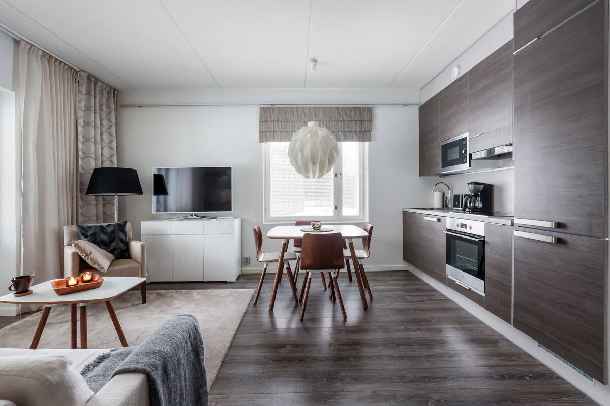 Una sala de estar moderna con un sofá, una mesa de centro y un televisor.  Junto a ella, un comedor con una mesa y sillas. La cocina cuenta con un microondas, horno y armarios de madera oscura.