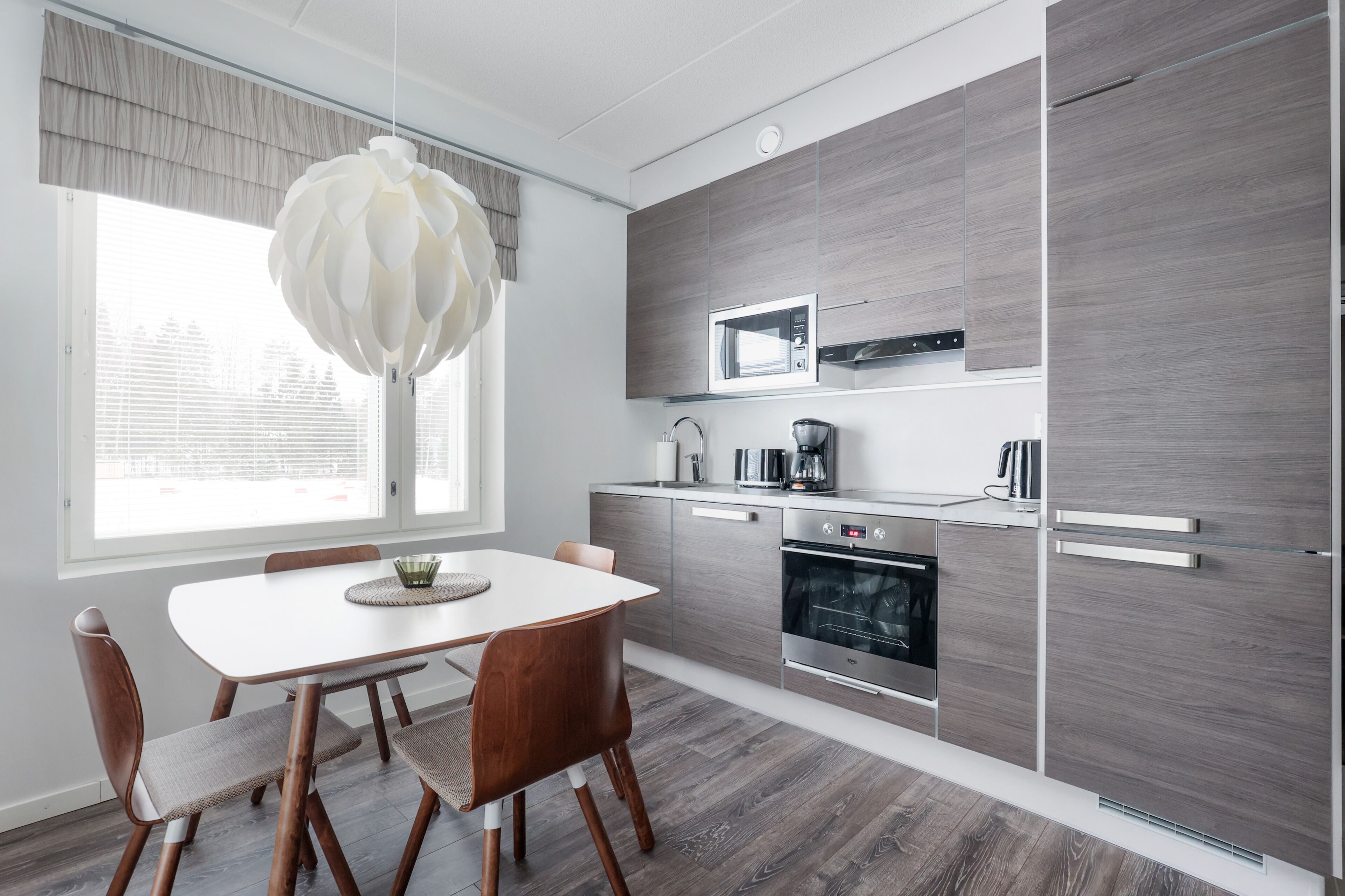 Een moderne keuken met een witte eettafel, houten stoelen, een hanglamp en een inbouwmagnetron.
