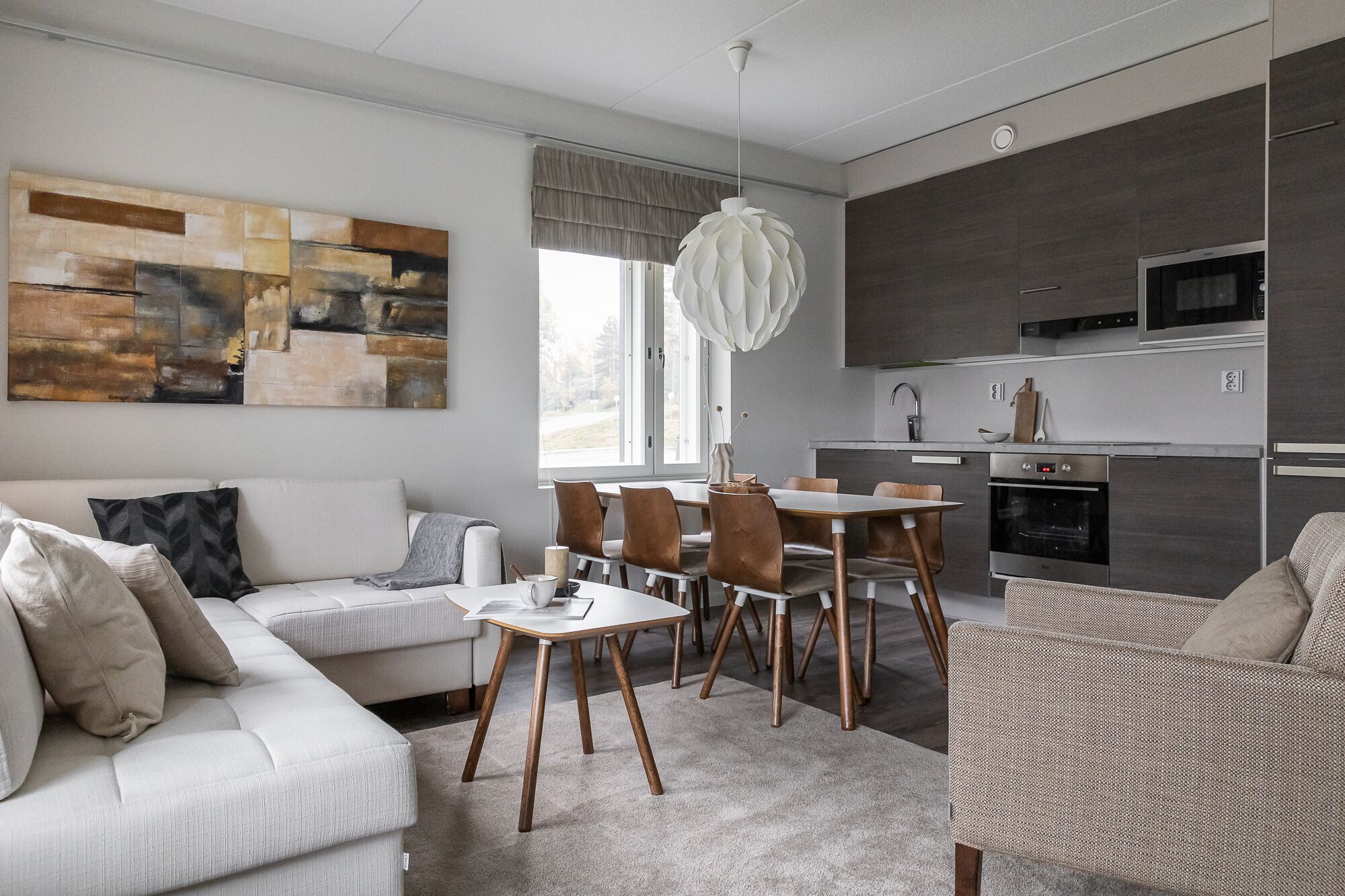 A modern living room with a sofa, dining table, and chairs. There is a kitchen area with appliances and a large window.