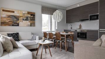 A modern living room with a sofa, dining table, and chairs. There is a kitchen area with appliances and a large window.