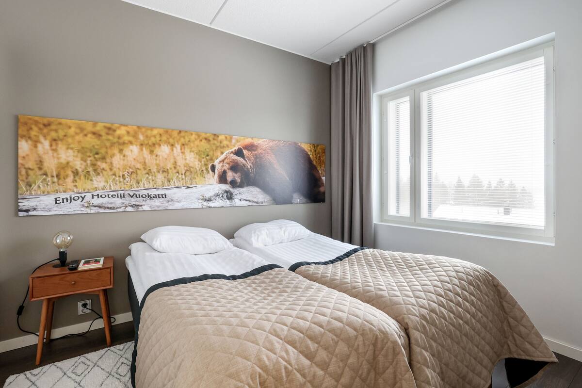 Una habitación de hotel con dos camas, una mesita de noche con lámpara, un cuadro de un oso y una ventana con persianas.
