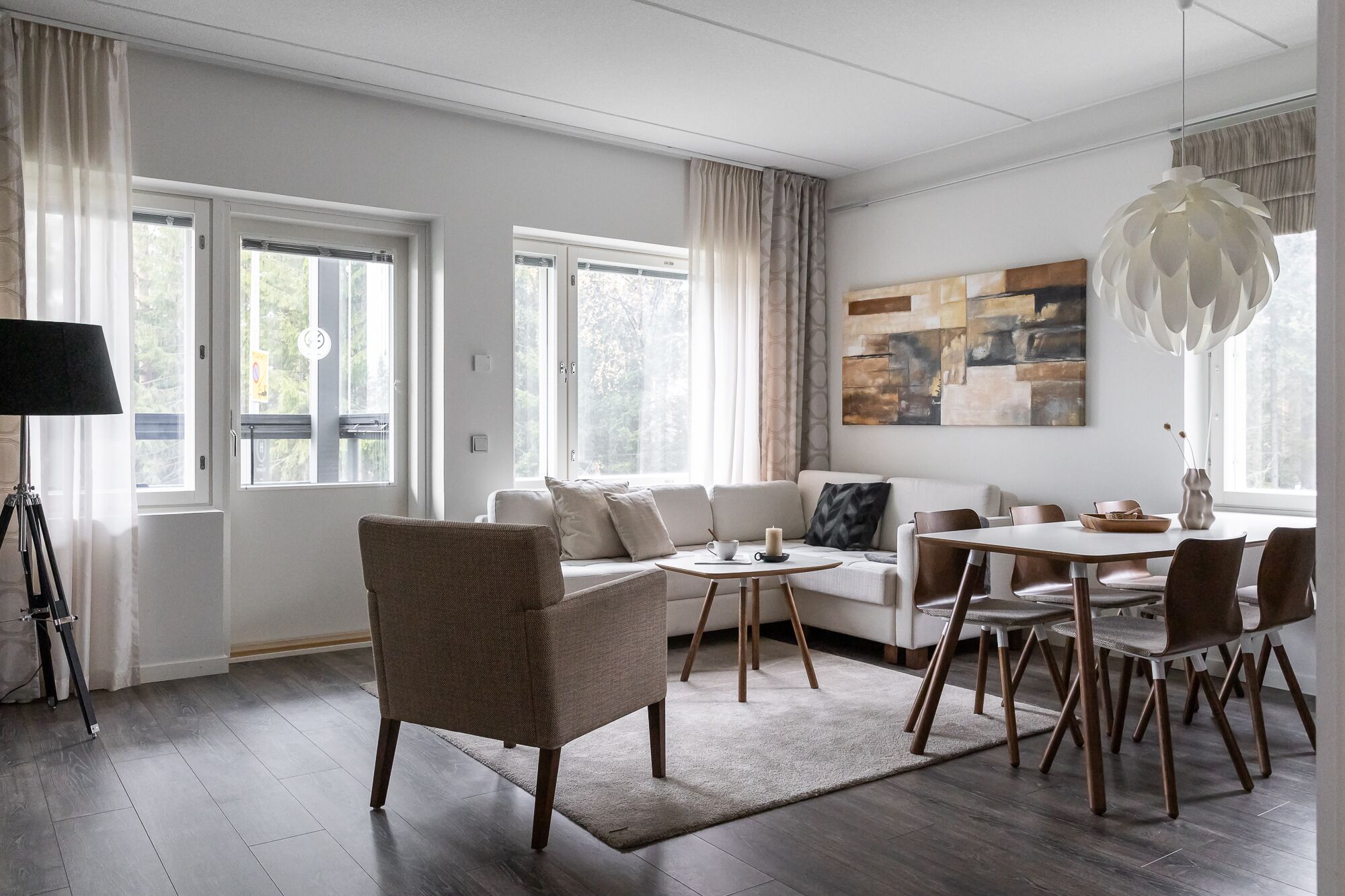 A modern living room with a sofa, dining table, and chairs. There are large windows with curtains, a lamp, and a painting on the wall.