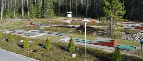 A miniature golf course with multiple green and red holes, surrounded by a forested area.