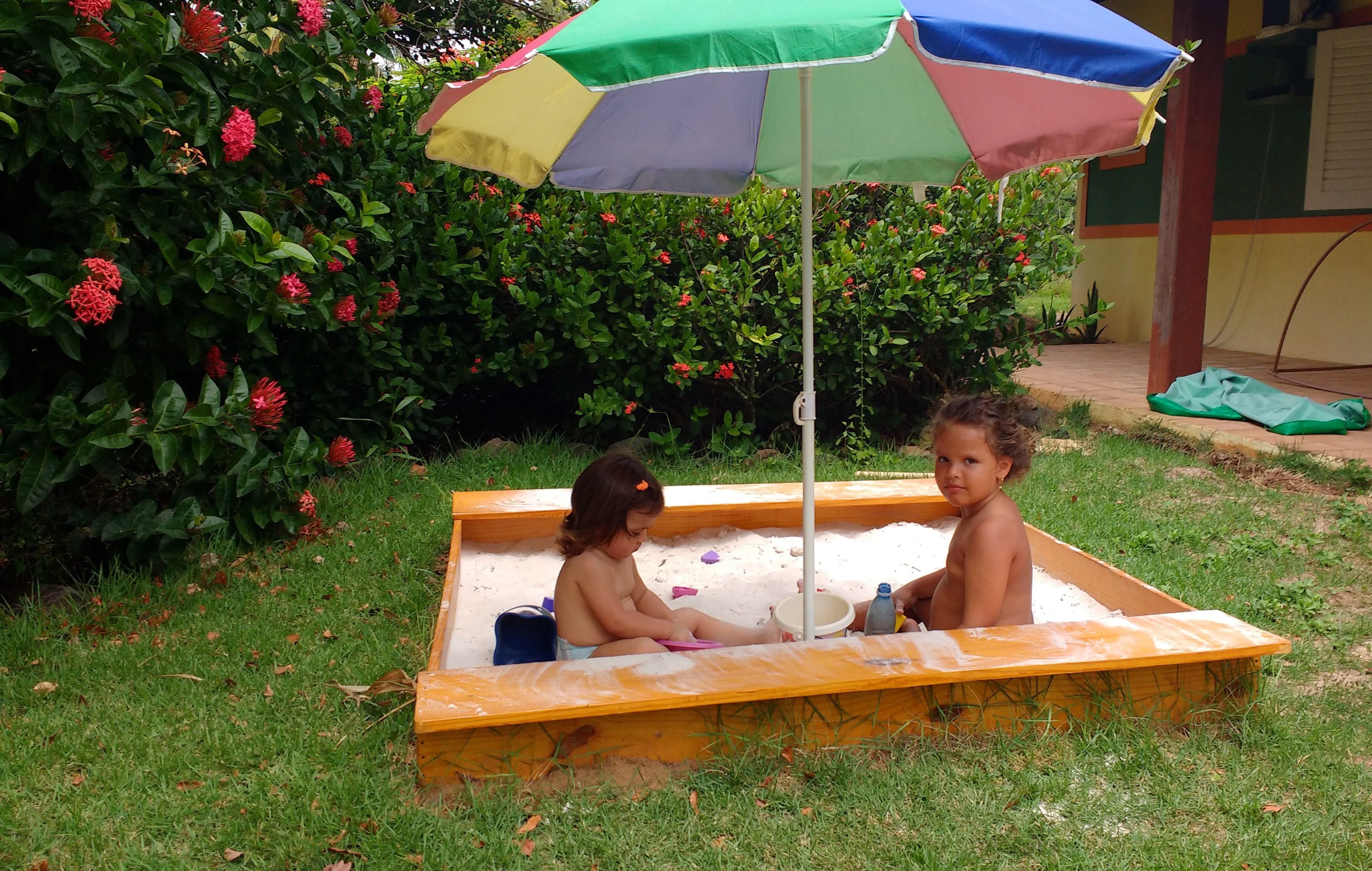 Dos niños jugando en un arenero bajo un paraguas colorido.