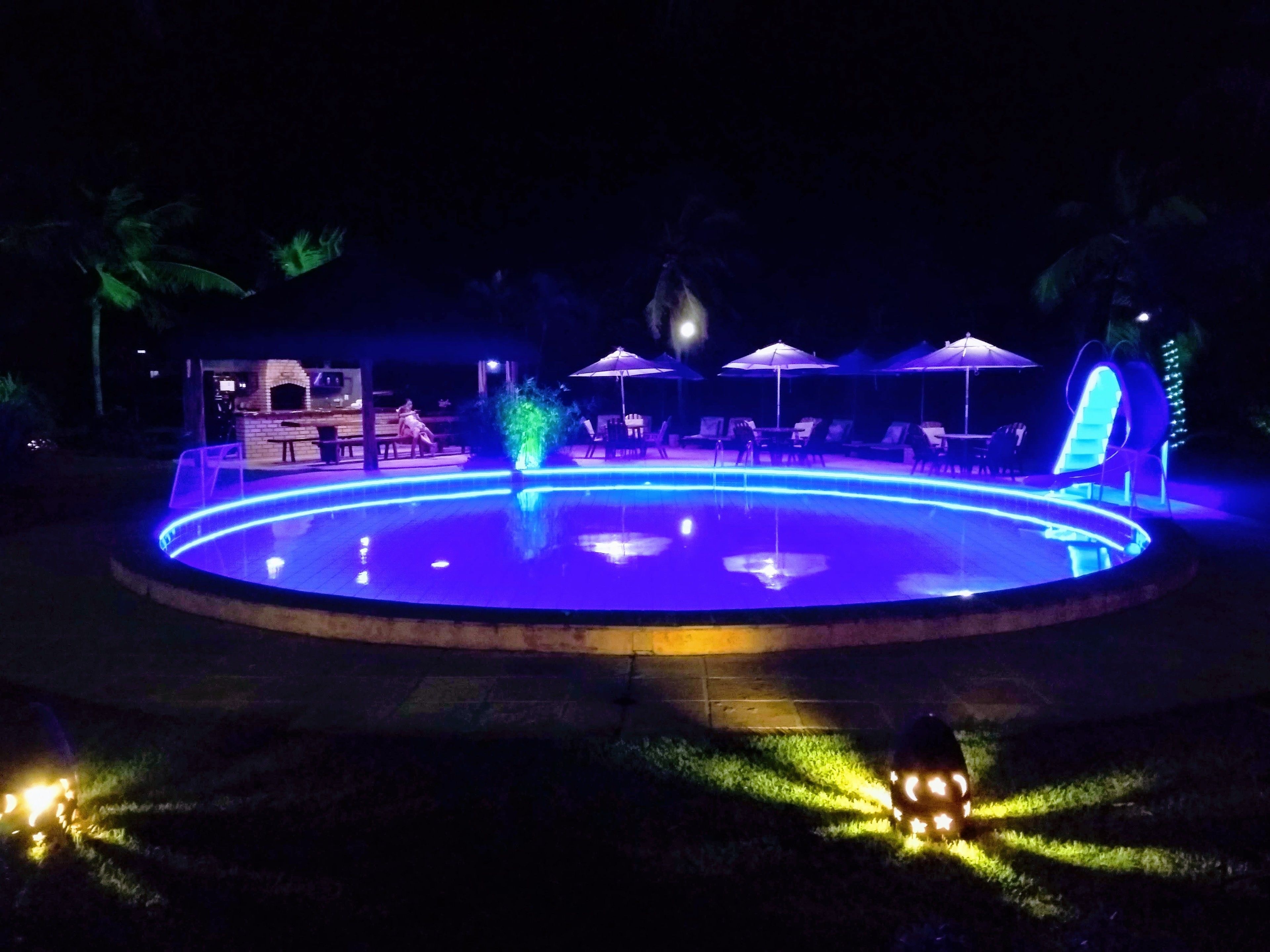 A circular pool with a fountain, surrounded by lounge chairs and umbrellas, and a small building in the background.